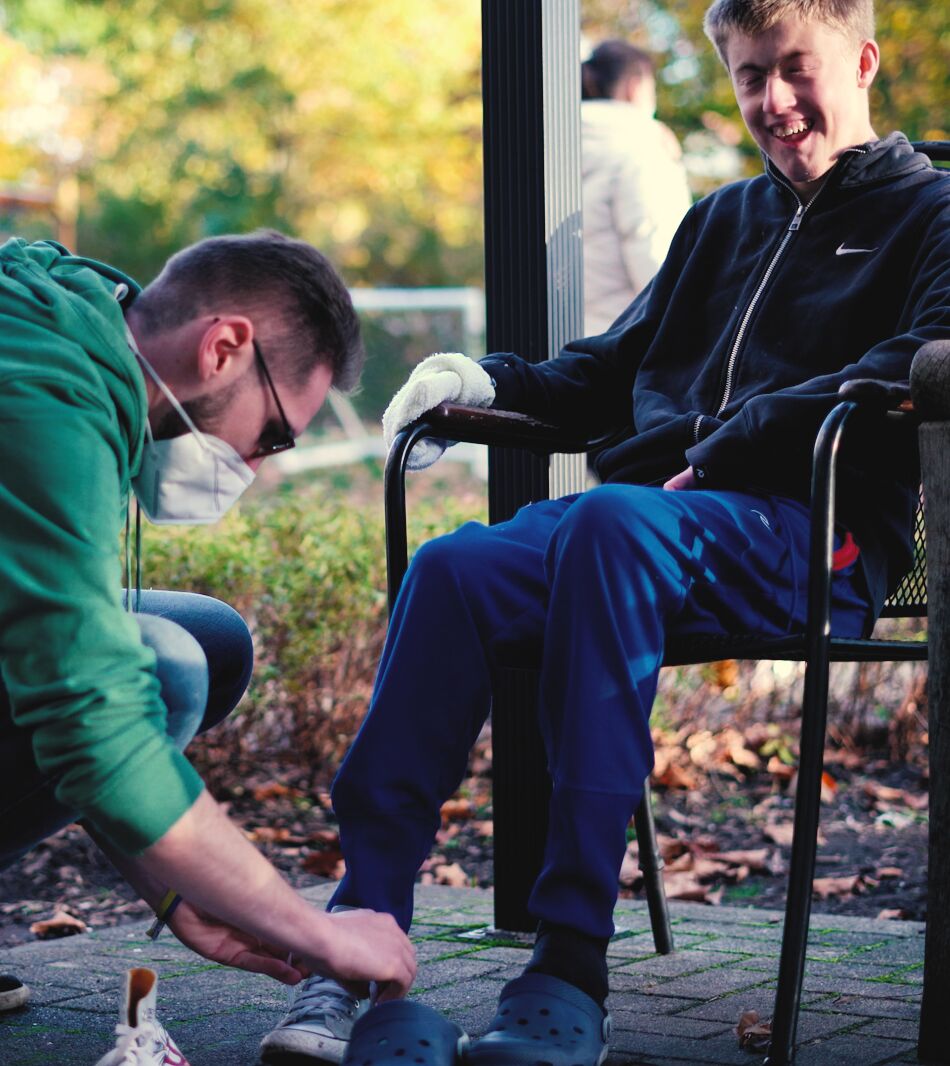 Ein Heilerziehungspfleger zieht einem lachenden Jungen die Schuhe an